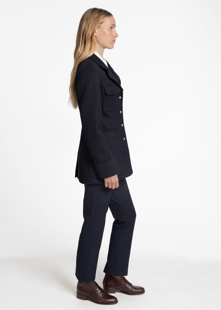 A woman in side profile wearing a dark navy jacket with flap chest pockets, matching navy trousers, and brown leather dress shoes, photographed against a clean white studio background.