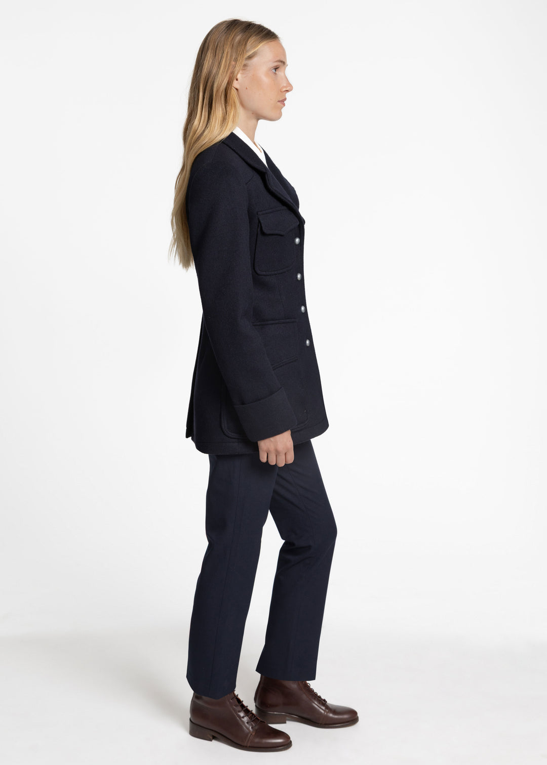 A woman in side profile wearing a dark navy jacket with flap chest pockets, matching navy trousers, and brown leather dress shoes, photographed against a clean white studio background.
