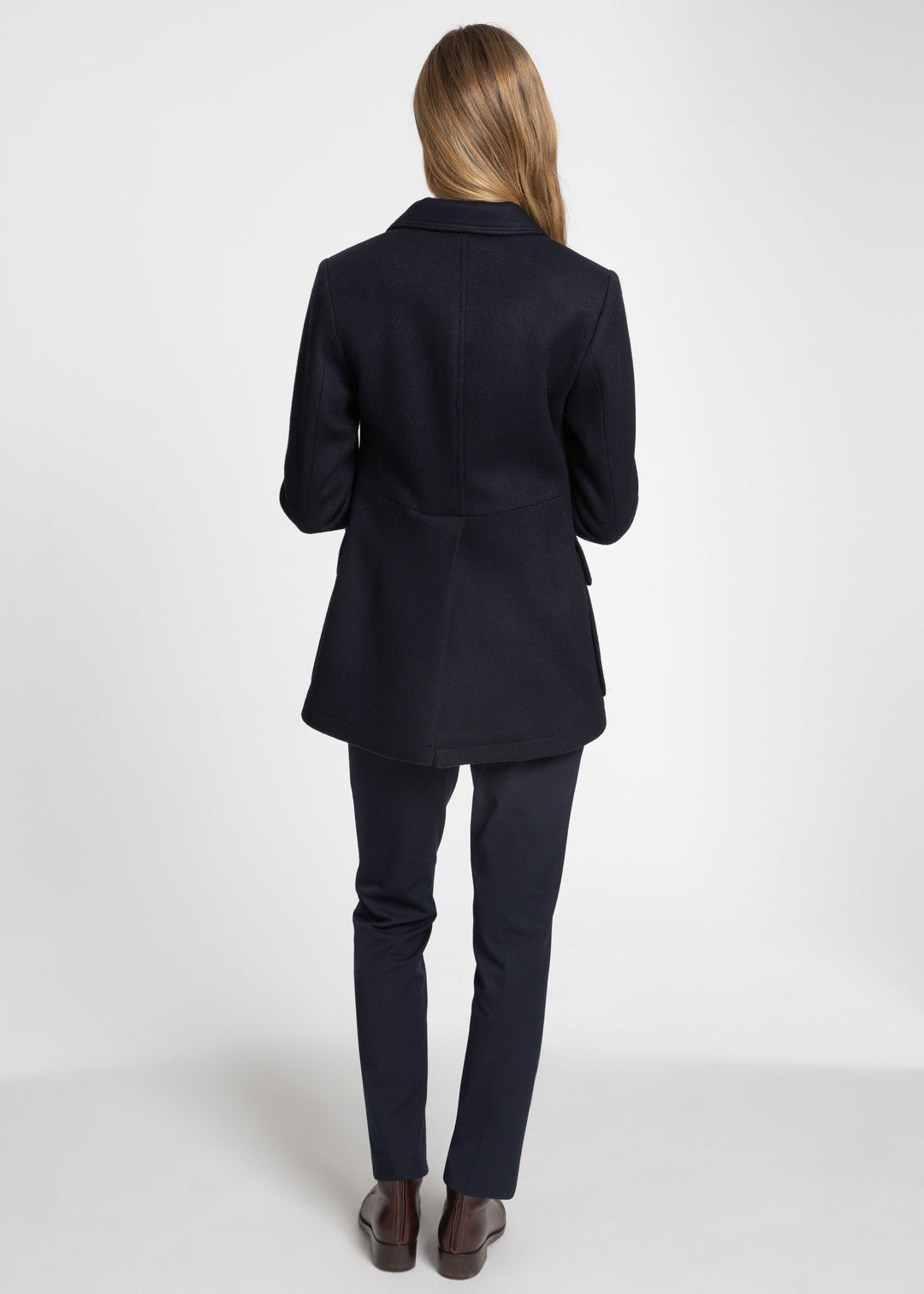 A woman shown from the back wearing a dark navy jacket and slim-fit trousers, standing against a clean light gray studio background.