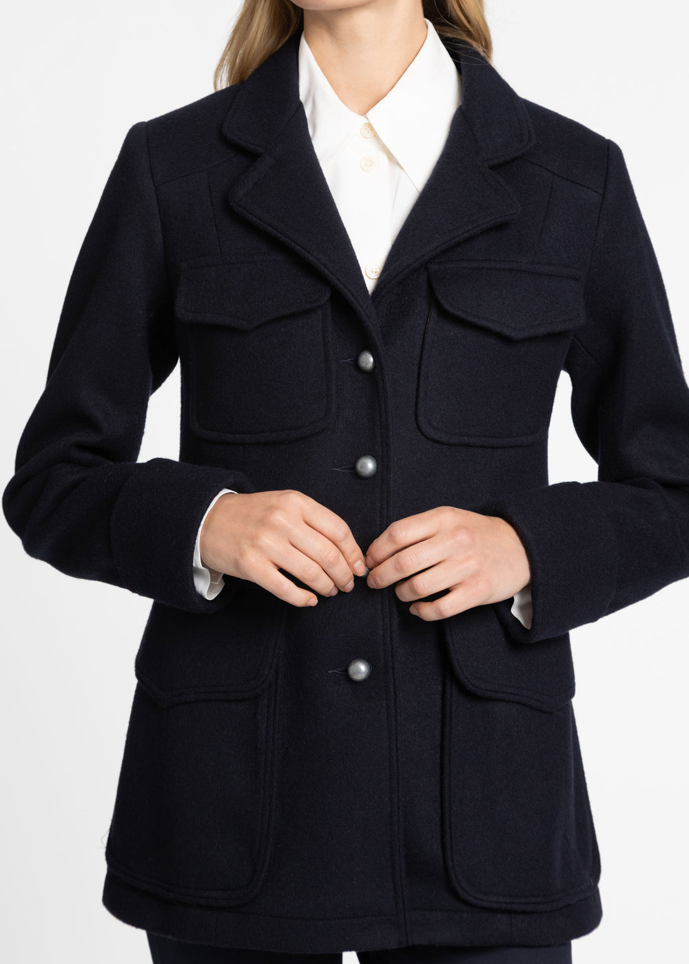 A woman wearing a dark navy wool-blend jacket with silver buttons, layered over a white collared shirt, shown from the neck down against a light gray studio background.