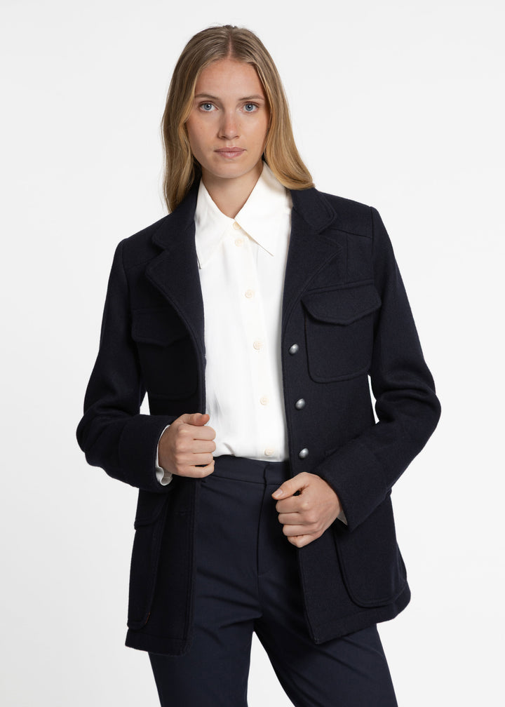 A woman modeling a dark navy jacket over a white collared blouse with matching navy trousers, photographed against a clean light gray studio background.