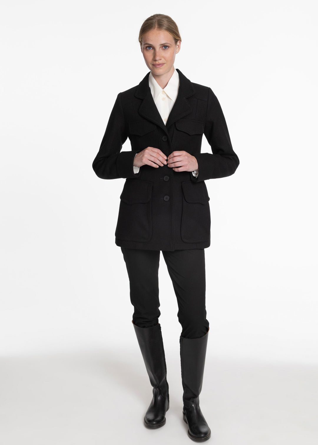 A woman modeling a black tailored jacket over a white collared shirt, black riding pants, and black riding boots against a plain white studio background.