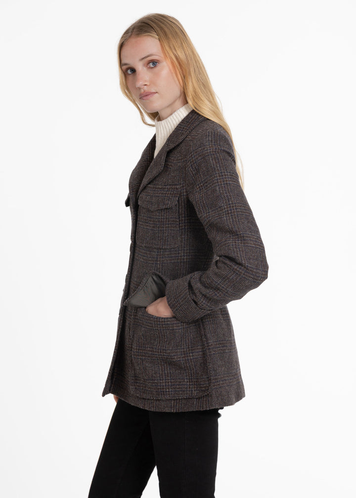 A woman modeling a dark brown and gray plaid blazer over a cream ribbed turtleneck and black pants, photographed against a plain white studio background.