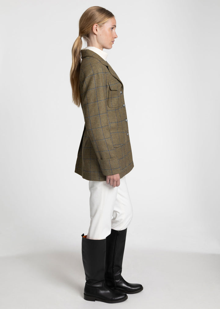 A woman shown in profile wearing a green plaid jacket, white riding breeches, and black riding boots against a light gray studio background.