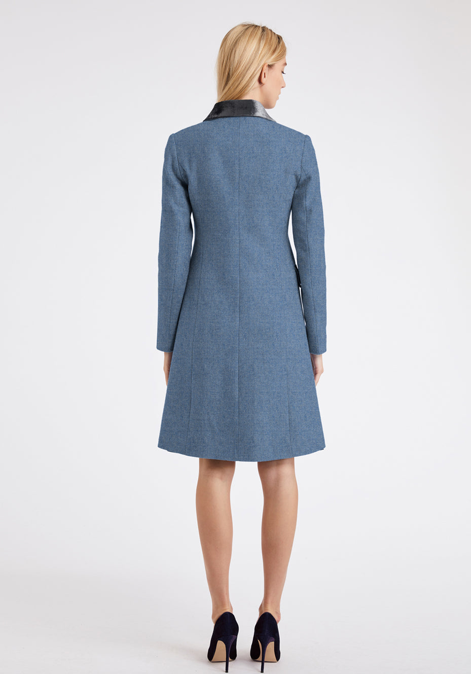 The back of a woman wearing a blue coat with a grey velvet collar trim on a white background