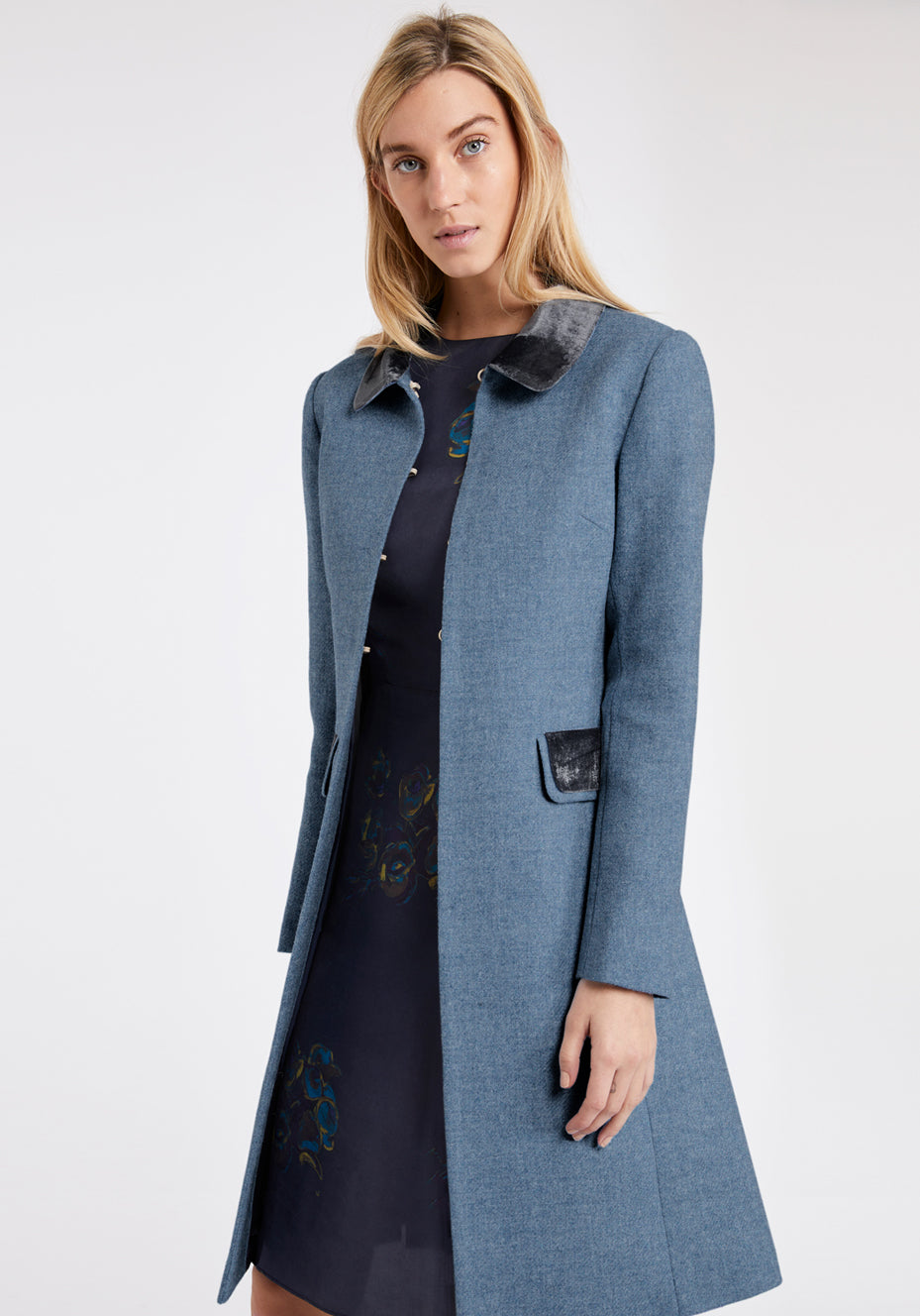 Woman wearing a long blue coat with velvet trim on the collar and pockets with a white background