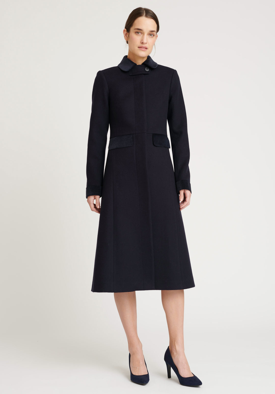 A model wears the Contrast Coat, a long and elegantly tailored navy wool coat with navy cotton velvet accents on the collar, cuffs, back belt, and pocket flaps.