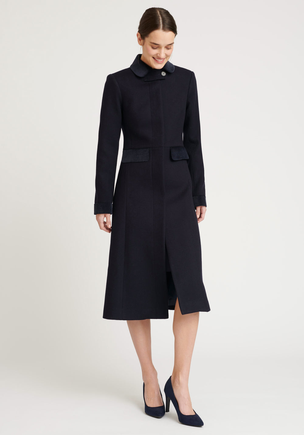 A model wears the Contrast Coat, a long and elegantly tailored navy wool coat with navy cotton velvet accents on the collar, cuffs, back belt, and pocket flaps.
