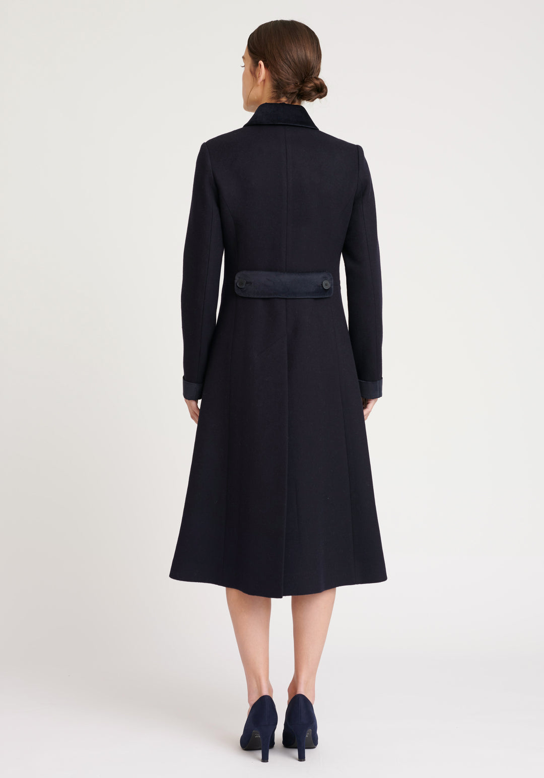 A model wears the Contrast Coat, a long and elegantly tailored navy wool coat with navy cotton velvet accents on the collar, cuffs, back belt, and pocket flaps.