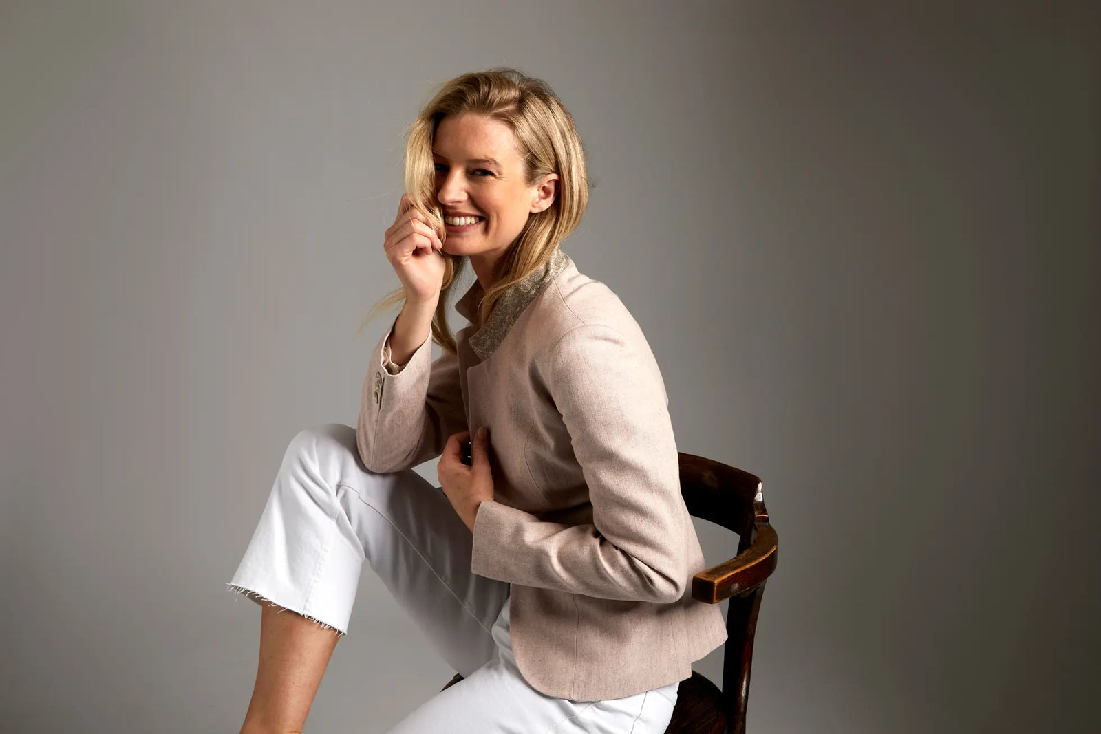 Woman sitting on a chair wearing a beige sweater and white pants against a gray background
