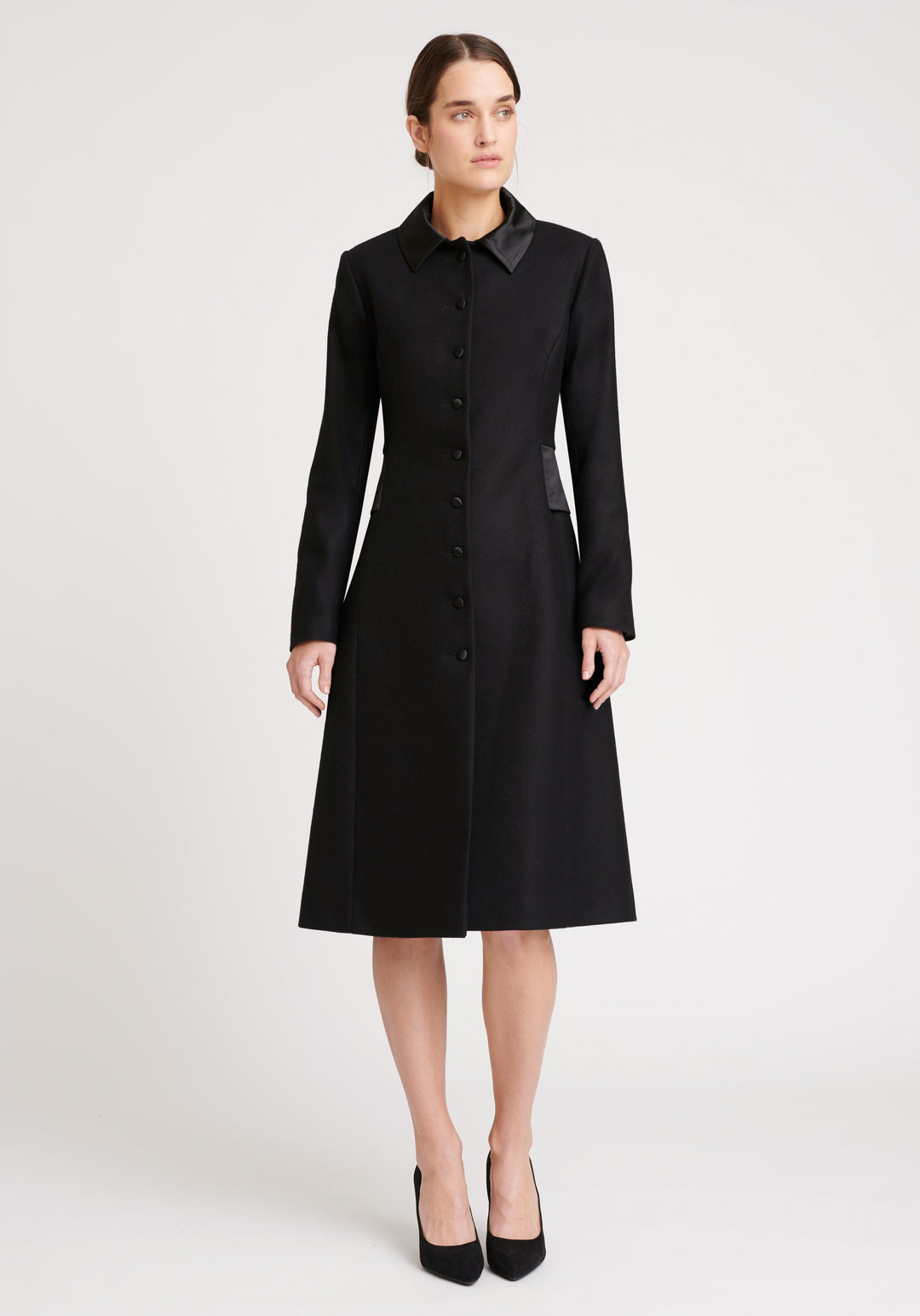 A model wears the Willa coat in black, an immaculately tailored piece with gently flared skirt and satin silk trim on the collar, side panels, and buttons.