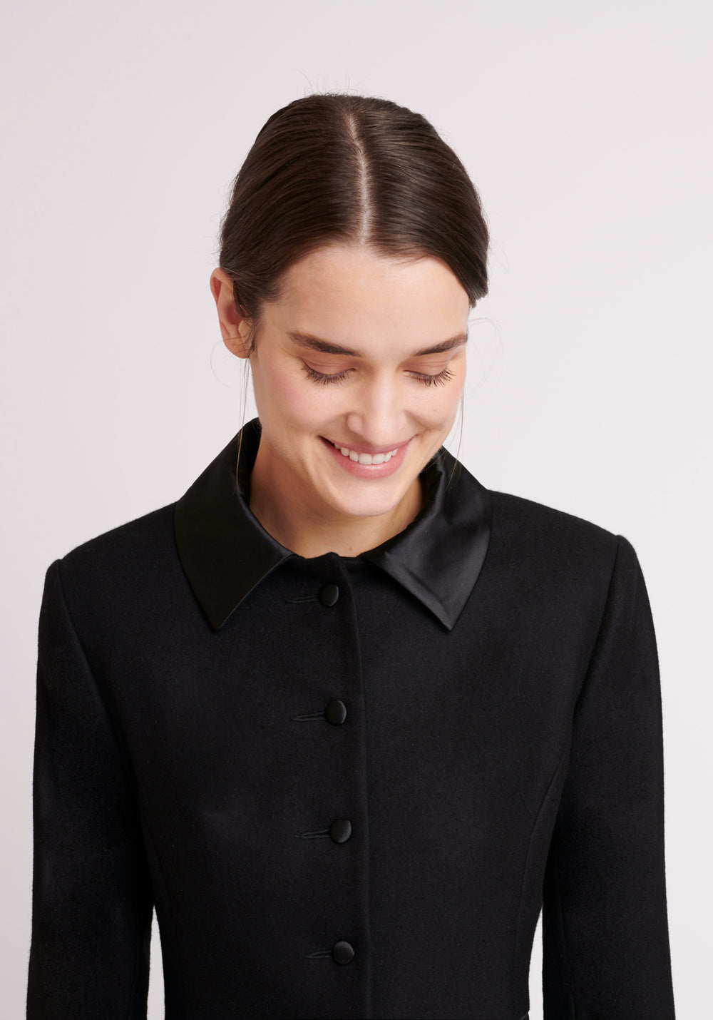 A model wears the Willa coat in black, an immaculately tailored piece with gently flared skirt and satin silk trim on the collar, side panels, and buttons.