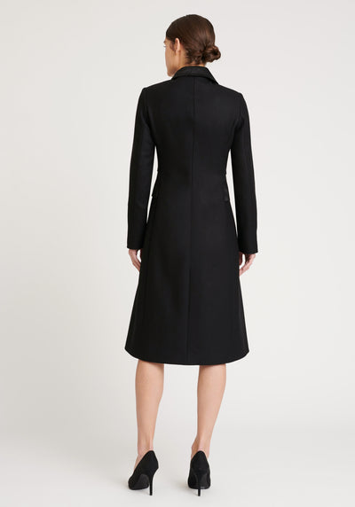 A model wears the Willa coat in black, an immaculately tailored piece with gently flared skirt and satin silk trim on the collar, side panels, and buttons.